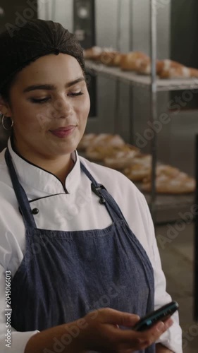 Vertical video: Checking phone baker in jacket and apron turning to greet customer at bakery POS