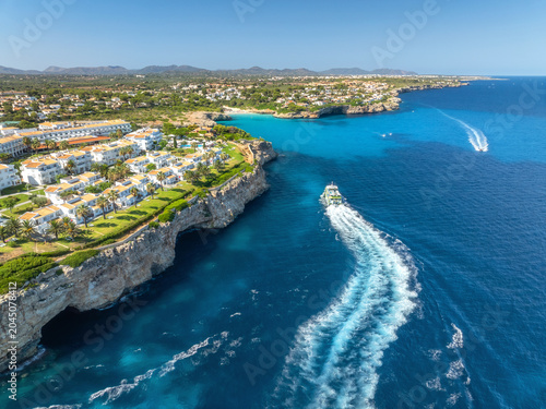Foto Aerial view of a white coastal resort with villas, palm trees on rocky cliffs above turquoise Mediterranean sea, boats on blue clear water