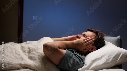 Fotografija Man lying awake in bed at night, clutching his face in pain and frustration from