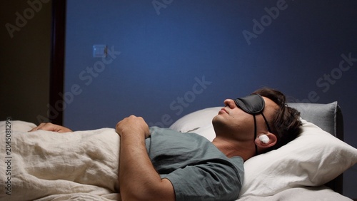 Konstfotografi Man sleeping peacefully in bed at night wearing an eye mask and earplugs for com