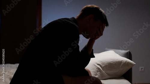 Foto Man sitting on the edge of a bed, covering his face with his hand, overwhelmed w