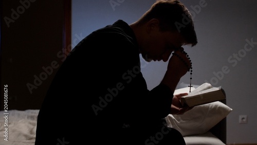 Εκτύπωση καμβά Young man seated in a dark room, clutching rosary beads and an open bible, eyes