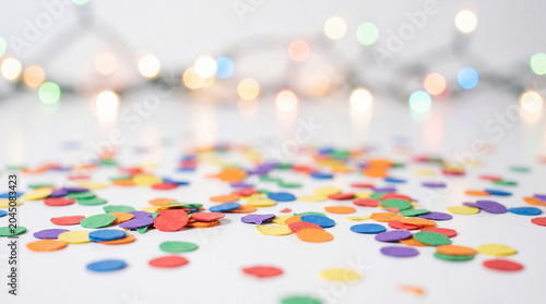 Colorful confetti scattered on white surface with blurred festive lights in background creating joyful and celebratory atmosphere
