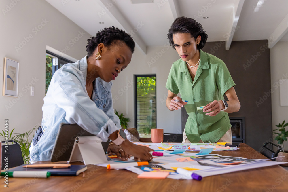 Obraz premium Diverse coworkers leaning and standing at table in home arranging layouts, tablet, sticky notes
