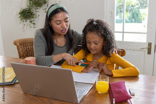 Diverse female adult and child sitting at dining table doing homework with laptop, pencils, juice