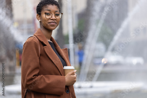 Foto African woman standing by fountain in plaza holding takeaway coffee cup, brown c