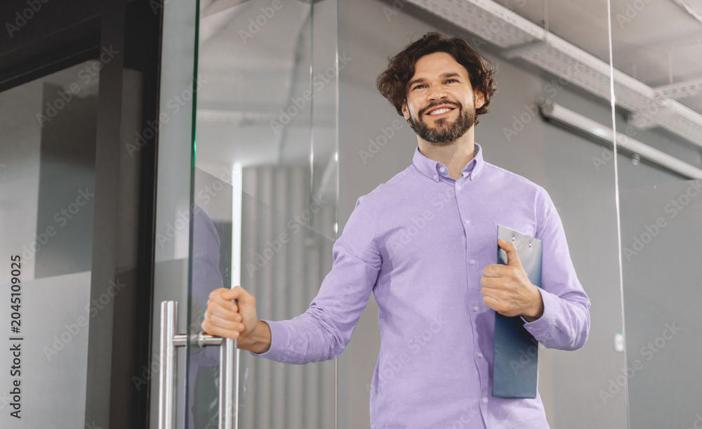 Fototapeta premium A man opens the glass door to a modern office. He smiles and holds a clipboard in one hand. The office features minimal design and bright lighting, suggesting a busy work environment