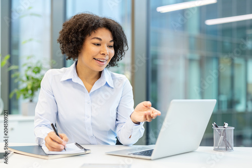 Smiling young African American woman is in the office, sitting at the table, talking on video call and taking notes