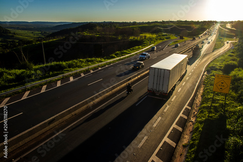 Highway SP-294 in Marilia, at sunset. Intense traffic of cars and trucks flows through the green landscape, illuminated by the golden hour light.