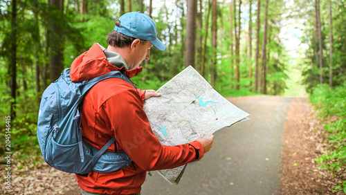 Slika na platnu Hiker looking at a map on a forest path, wearing a orange waterproof jacket and backpack, holding a paper map with trails