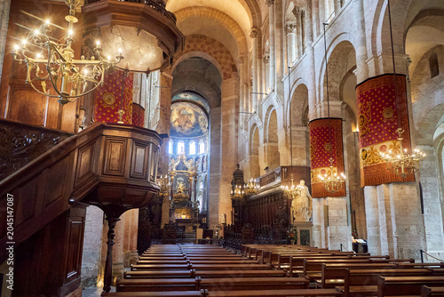 Bild auf Leinwand Interior of a Romanesque cathedral features vaulted ceilings, stone columns, red tapestries, ornate chandeliers, and wooden pews under soft natural light; cathedral interior