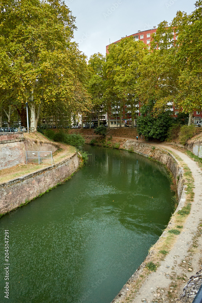 Fototapeta premium A narrow, tree-lined canal curves through a quiet urban area with stone embankments and a dirt path. Buildings rise behind dense green foliage; canal.