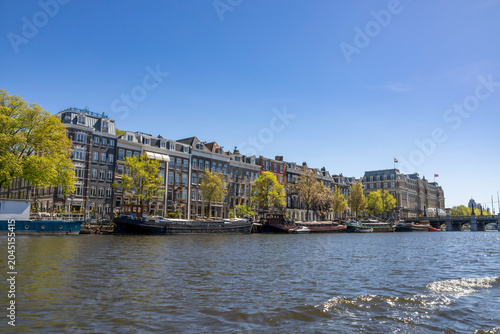Scenic view of Amsterdam's historic buildings along a picturesque canal under a bright blue sky, showcasing the city's unique architecture and tranquil waterways.