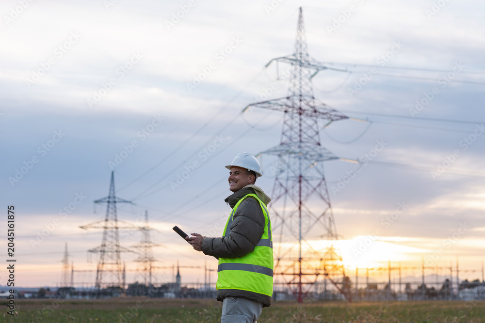 Obraz premium An electrical engineer and utility worker checks data on a tablet near high voltage power lines in the evening.