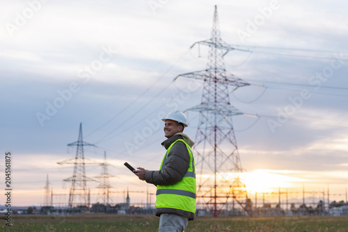 An electrical engineer and utility worker checks data on a tablet near high voltage power lines in the evening.