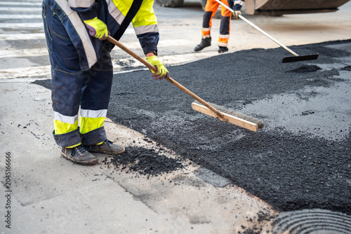 Sidewalk maintenance in urban city, workers evenly distribute asphalt mixture on road section with brushes and preparing surface for compacting new asphalt with roller machine, paving process in town