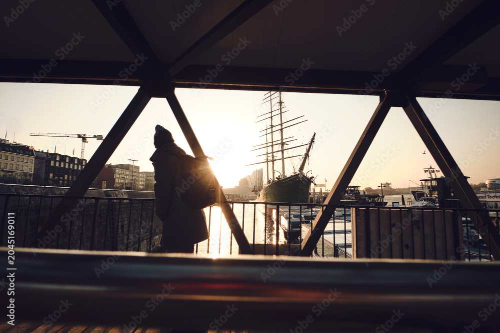 Naklejka premium A lone figure stands on a metal-framed pedestrian bridge overlooking a harbor at sunset. Backlit by the low sun, the person is rendered as a dark silhouette