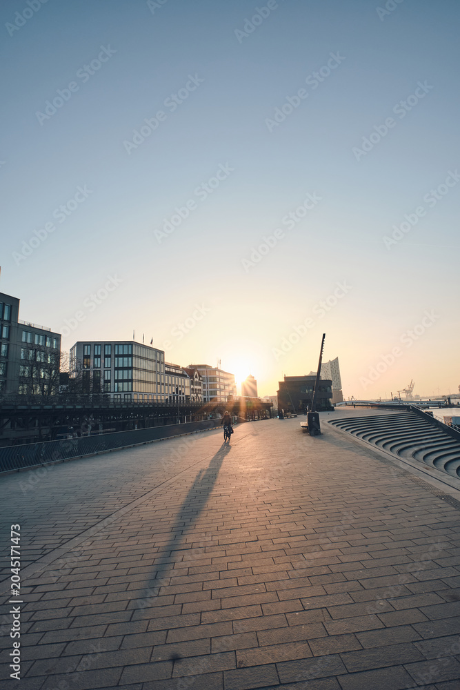 Obraz premium A wide paved promenade beside a city harbor at sunset, with low sunlight casting long silhouettes of a lone cyclist and distant pedestrians. Modern waterfront