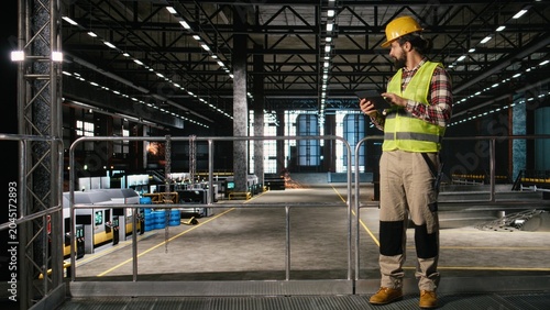 Fotografía Man engineer wearing helmet and checking production line on tablet, walking through industrial space filled with massive machinery, Warehouse environment showcases heavy industry worksite