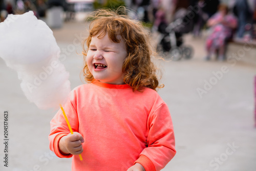 Ginger kid eating white cotton candy. Curly-haired child enjoys sugary snack in public park. Summertime treat for toddler experience, sweet dessert fun concept.