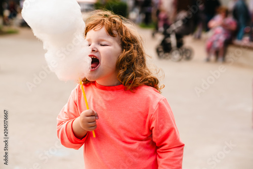 Ginger kid eating white cotton candy. Curly-haired child enjoys sugary snack in public park. Summertime treat for toddler experience, sweet dessert fun concept.