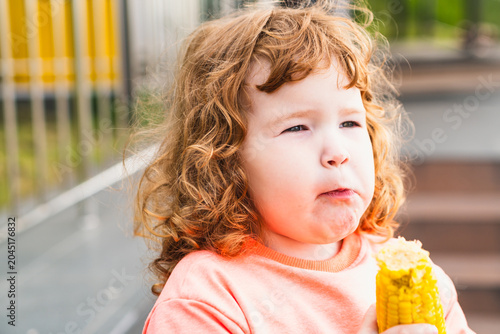 Toddler girl eating boiled yellow corn cob outdoors. Cute ginger kid with curly hair holding healthy snack. Childhood lifestyle, organic nutrition, vegetable diet and summer food concept.