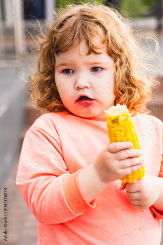 Toddler girl eating boiled yellow corn cob outdoors. Cute ginger kid with curly hair holding healthy snack. Childhood lifestyle, organic nutrition, vegetable diet and summer food concept.
