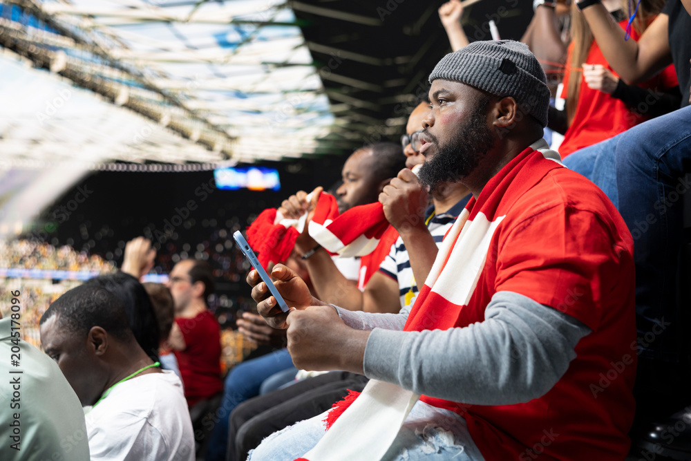 Naklejka premium Black fan does live betting during football game at the stadium filled with anticipation and excitement. Supporter check live odds on smartphone, tension and high risk of sports betting live.
