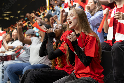 Photos Young female football supporter holding red scarf near her mouth, with face paint anxiously watching tense live soccer match from stadium stands