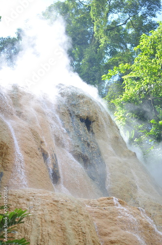 A waterfall with steam rising from it. The steam is white and the water is brown. The waterfall is surrounded by trees