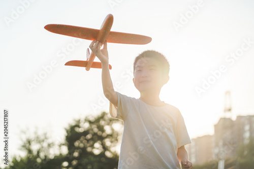Cuadro en lienzo A young Asian boy holds an orange toy glider airplane aloft while playing outdoors during golden hour sunset, with warm backlighting filtering over him