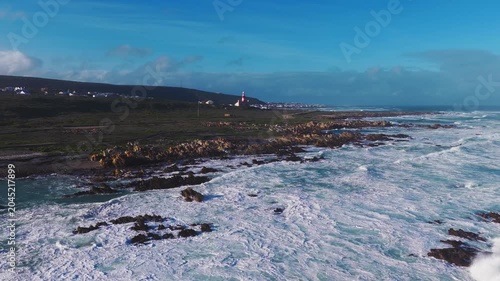 Aerial view glides parallel to the shore at Cape Agulhas, South Africa. Powerful surf breaks by dark rocks, turquoise water, and a red and white lighthouse in clear daylight.