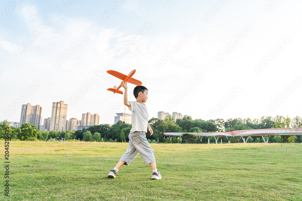 Obraz premium An East Asian young boy holds an orange toy airplane while walking on a spacious grassy park field under a bright daytime sky.