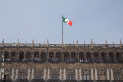 A majestic historic building featuring ornate colonial architecture with the Mexican national flag waving above the rooftop against a clear blue sky