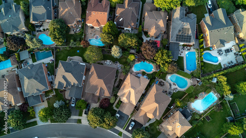 A high top-down aerial drone photograph showing a bird's-eye view of a row of upscale suburban houses, manicured green lawns, mature trees, and multiple backyard swimming pools in summer.