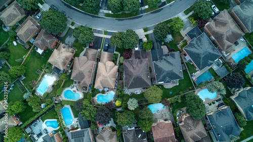 Photos A direct overhead drone photograph capturing a row of residential suburban homes situated along a curved street cul-de-sac, featuring multiple backyard swimming pools and manicured green lawns