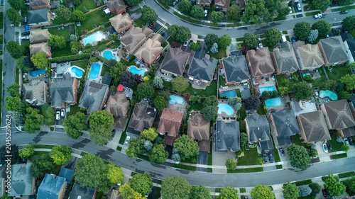 Quadro su tela A high-altitude aerial drone photograph looking straight down at a sprawling suburban development featuring winding, curved asphalt streets lined with detached family homes and backyard pools