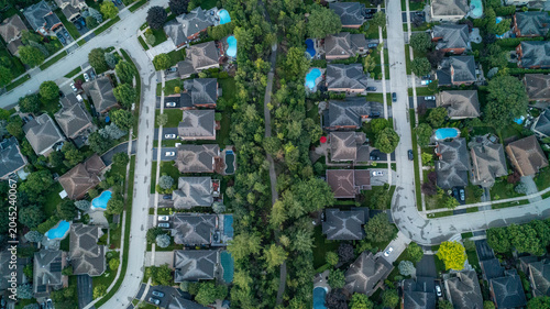 A vertical high-angle drone photograph looking down at two rows of suburban residential homes and backyard swimming pools divided down the center by a dense green belt of trees and a walking trail.