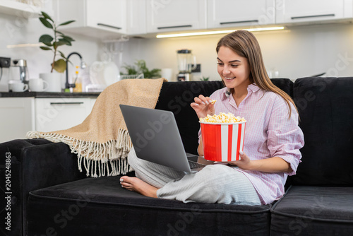 Canvas Print Young woman at home streams comedy on laptop eats popcorn and laughs out loud with happy mood