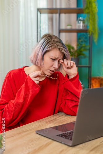 Young woman at home laptop removes glasses rubbing eyes suffering screen fatigue after remote work shift. Girl with pastel hair in red shirt closes eyes feeling headache seeking break for vision care