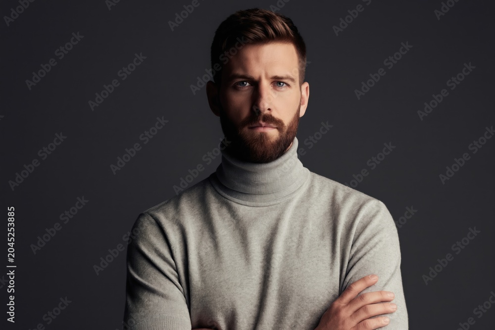 Obraz premium Studio portrait of bearded man with folded arms. He wears grey turtleneck sweater. His gaze is direct and serious, conveying professionalism and confidence, suitable for business or lifestyle themes.