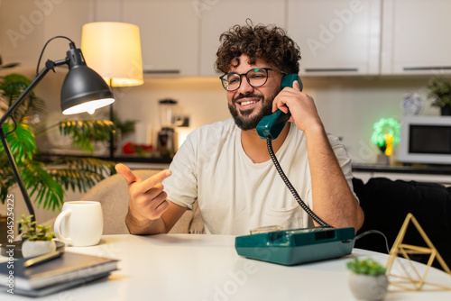 Arabian man at home on hotline talks on corded telephone listens asks brief questions solves issue. Egyptian guy sitting at table writes ticket on laptop gives steps polite voice calm support tonight.