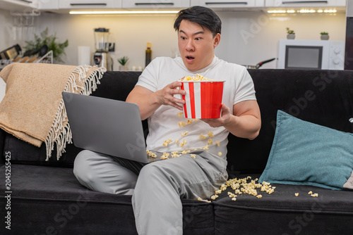 Photos Asian man at home streams tense film on laptop flinches at jump scare drops popcorn steadies breath