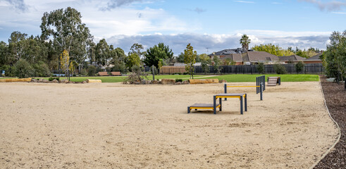 A spacious off-leash dog park set within a suburban reserve, featuring sandy ground, agility equipment, eucalyptus trees border the fenced enclosure, residential homes nearby in Melbourne Australia