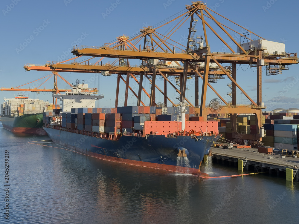 Naklejka premium container ship with water from anchor moored at container terminal. cargo operation in progress. big overhead type cranes working