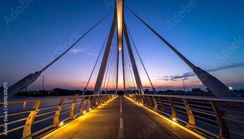 Futuristic Pedestrian Bridge at Dusk: A modern, illuminated pedestrian bridge stretches across the water at twilight, its sleek cables and warm lights creating a captivating urban landscape.