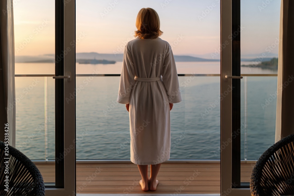 Fototapeta premium Woman in bathrobe on balcony at sunset overlooking calm ocean, luxury relaxation vacation