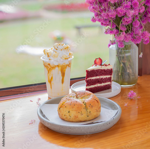 A tempting bakery set featuring a cream cheese garlic bread bun, a slice of red velvet cake, and an iced caramel popcorn coffee placed on a wooden counter by a cafe window.