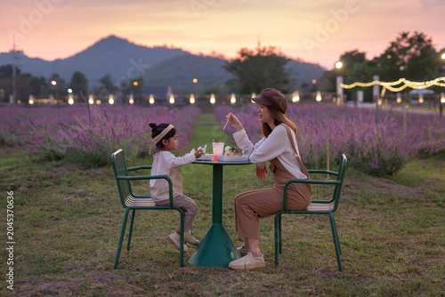 A heartwarming scene of a young Asian in brown overalls and her cute toddler daughter sitting at a round green table, eating dessert  in a blooming purple flower field at sunset.