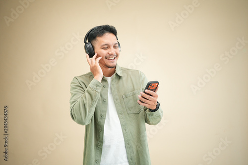 Smiling young Asian man holding smartphone and adjusting wireless headphones, looking at phone with casual expression in studio portrait